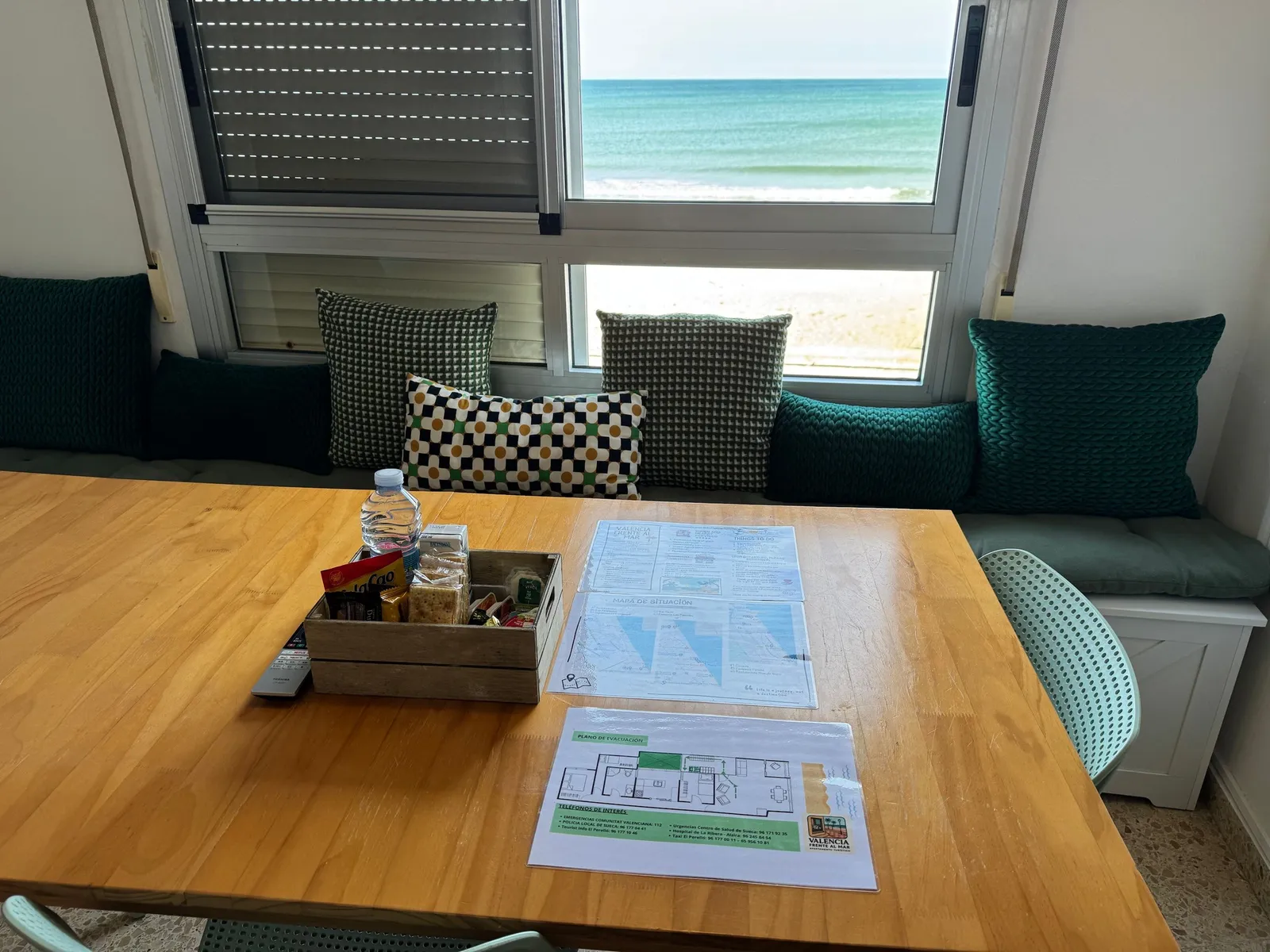 Mesa del salón junto al ventanal, con el mar de fondo, la guía del apartamento y una caja de bienvenida