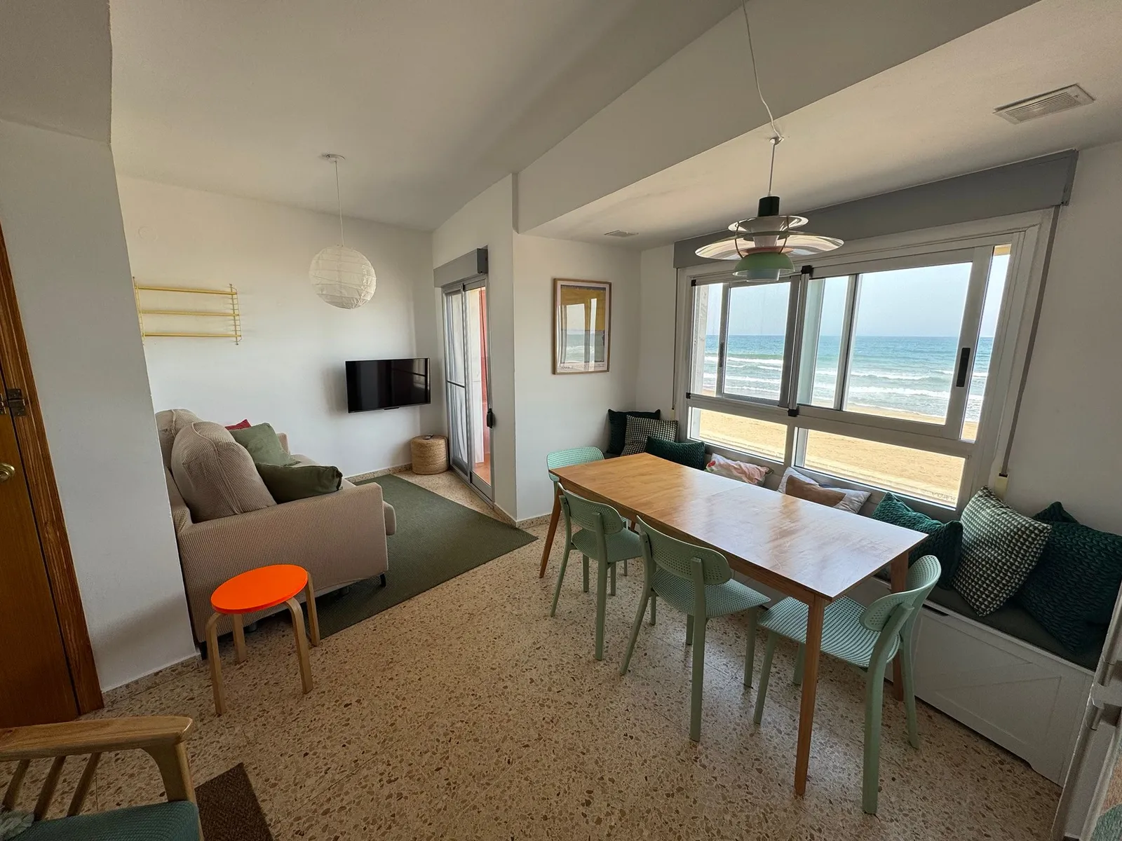 Salón-comedor con mesa de madera, banco corrido con cojines junto al ventanal y la playa de la Llastra al otro lado del cristal